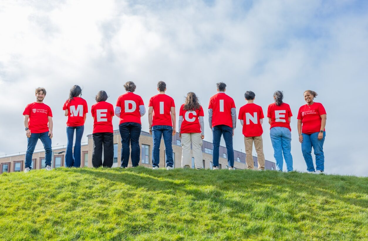 Students standing on a hill above the medical school, wearing red tee shirts, on the back of which each has a letter spelling Medicine